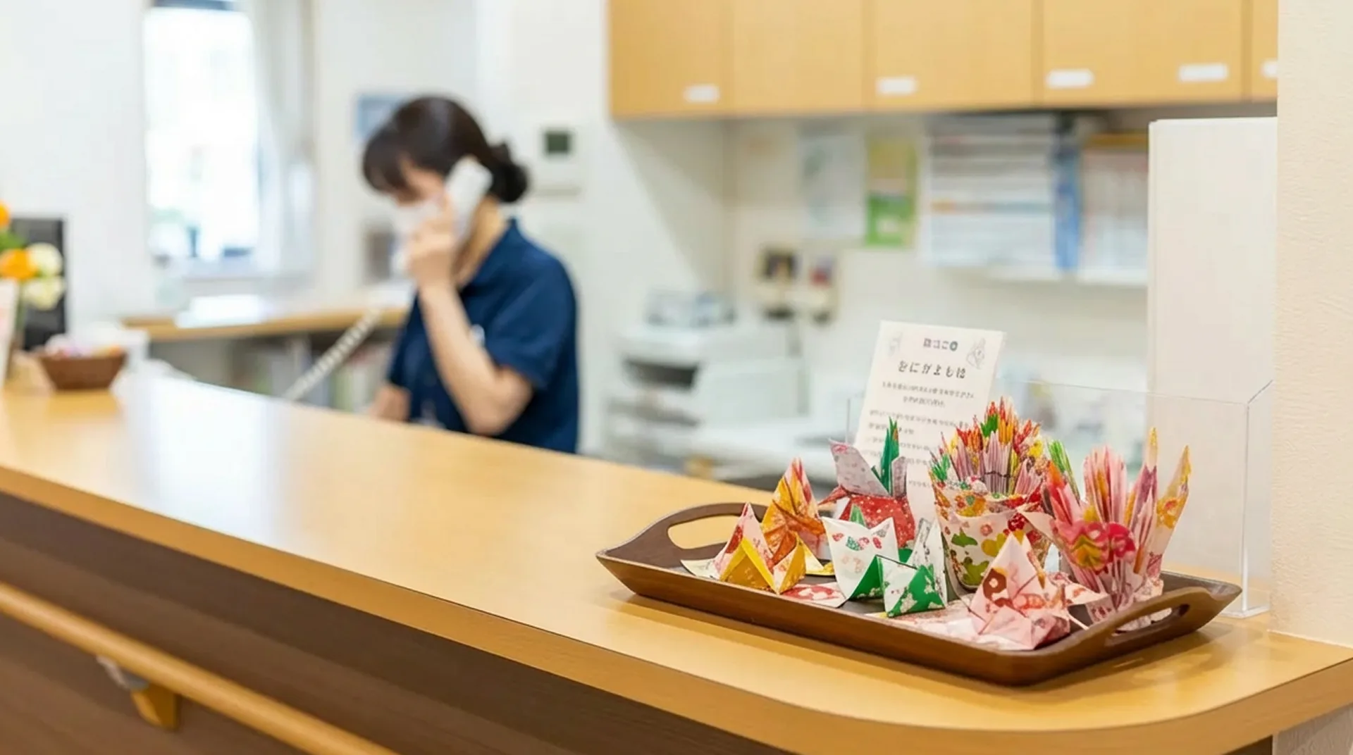 受付で電話対応する女性スタッフと飾り物 受付カウンターで電話対応をしている女性スタッフが写っています。手前には折り紙で作られたカラフルな飾り物がトレイに置かれており、明るく温かみのある雰囲気です。